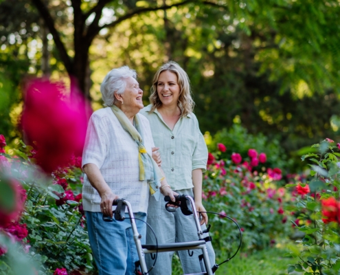 elderly woman using a walker and a younger woman walking beside her in a garden filled with vibrant flowers, including red roses. Both women are smiling and appear to be enjoying each other's company. The setting is outdoors with lush green foliage and trees in the background, suggesting a peaceful and pleasant environment. The elderly woman is wearing a light-colored blouse with a scarf, while the younger woman is dressed in a light green shirt and pants. The composition highlights the beauty of nature and the warmth of their relationship.