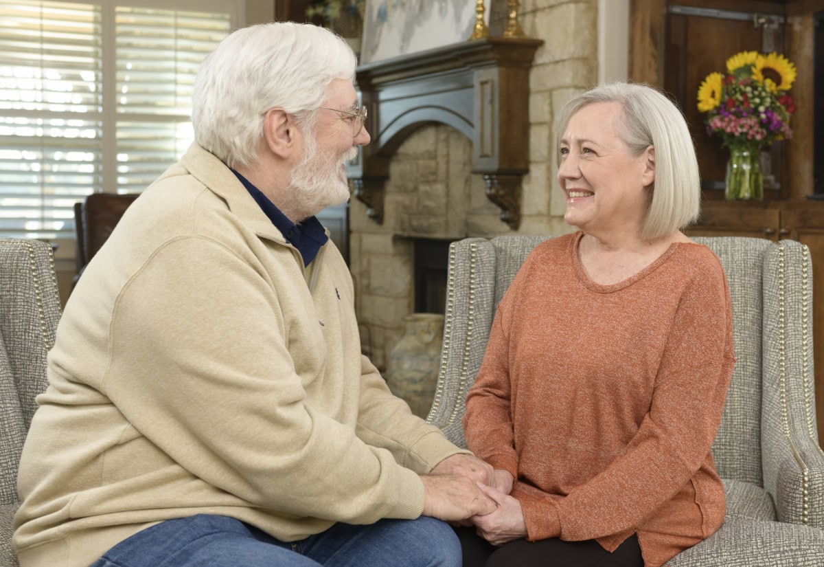 Elderly man and woman talking and smiling in a cozy living room.